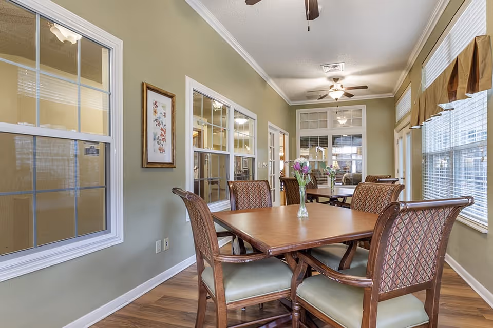 A dining area with wooden tables and cushioned chairs arranged neatly. Each table has a small vase with flowers. The room has light green walls, large windows with blinds and valances, and ceiling fans with lights. There are framed pictures on the walls and windows looking into adjacent rooms.