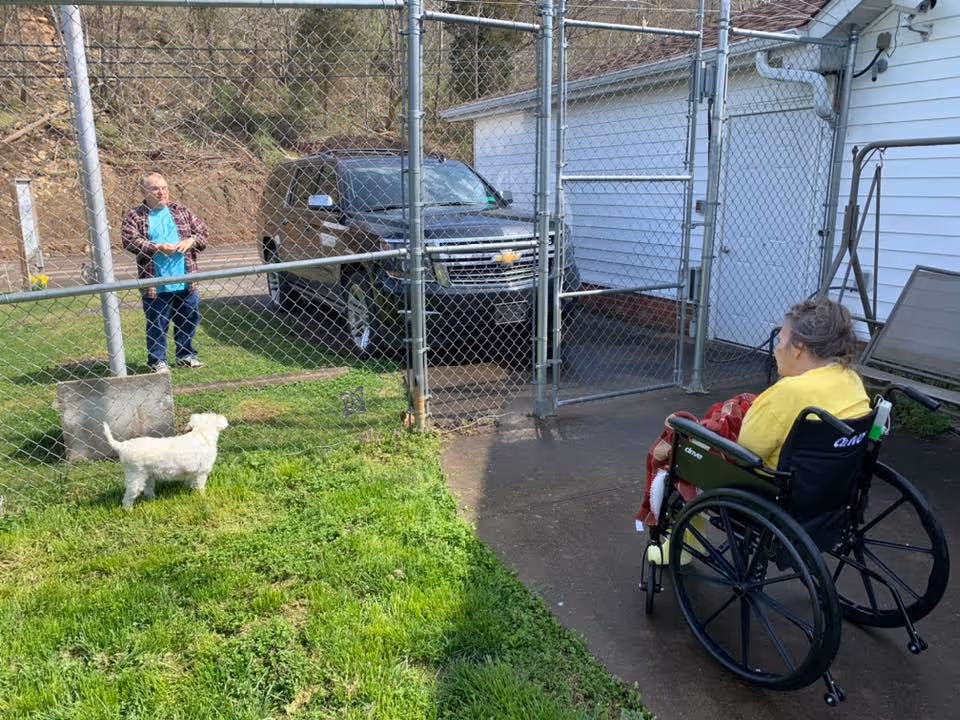 An elderly woman in a wheelchair on a concrete patio looks through a chain-link fence at an elderly man standing on grass with a small white dog. Behind the man is a black SUV parked near a white building.