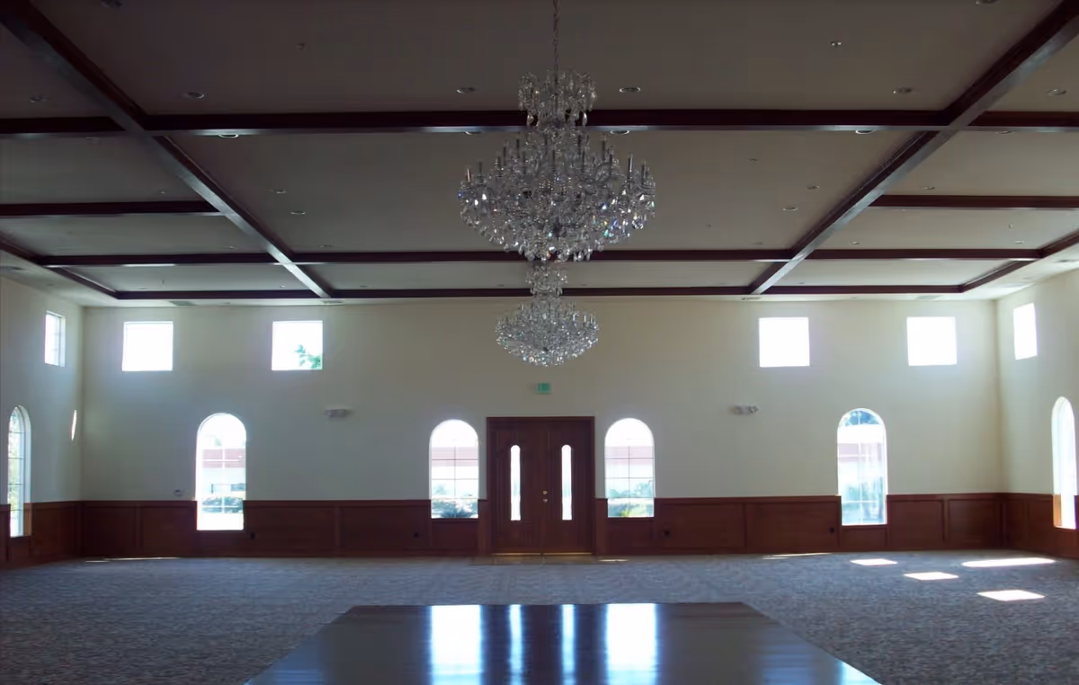 Large empty banquet-style hall with two crystal chandeliers, a coffered ceiling, arched windows and double doors.