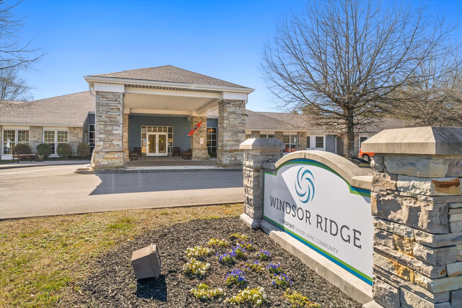 Exterior view of the Windsor Ridge senior living community building with a stone entrance and a sign in the foreground displaying the facility's name. The sky is clear and blue, and there are some trees and flowers around the sign.