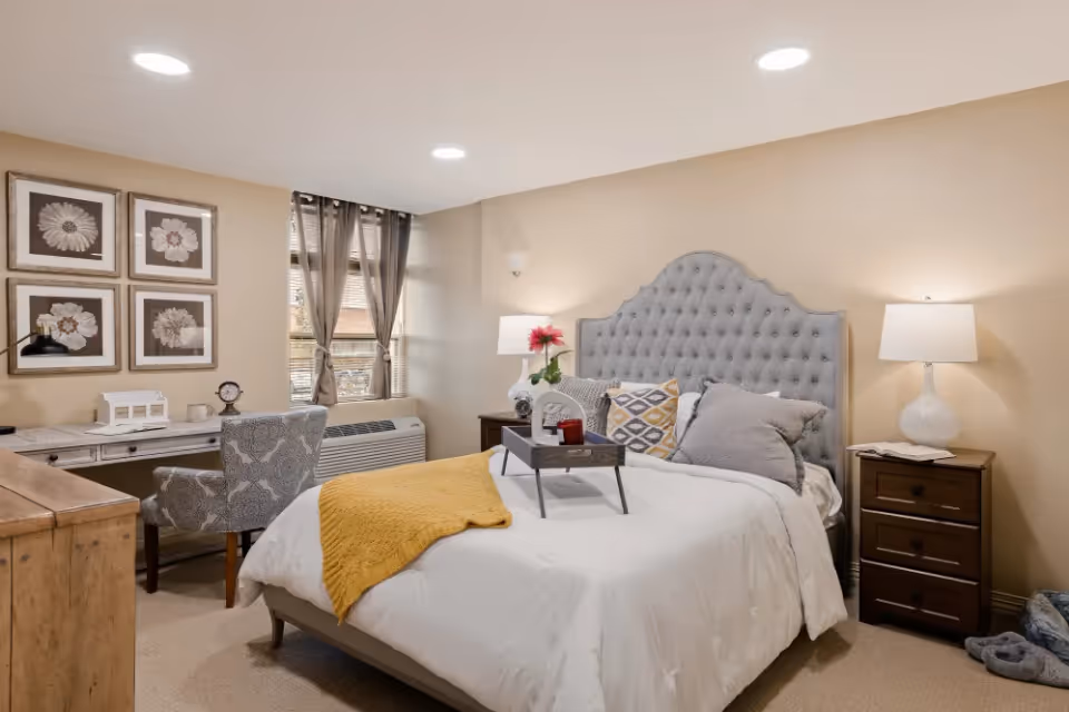 A cozy bedroom with a tufted gray headboard bed dressed in white bedding and a yellow throw blanket. There are multiple pillows on the bed and a small tray with a flower vase and candle. The room has beige walls, two bedside tables with lamps, a wooden dresser, a desk with a patterned chair, and four framed flower pictures on the wall. A window with sheer curtains lets in natural light.