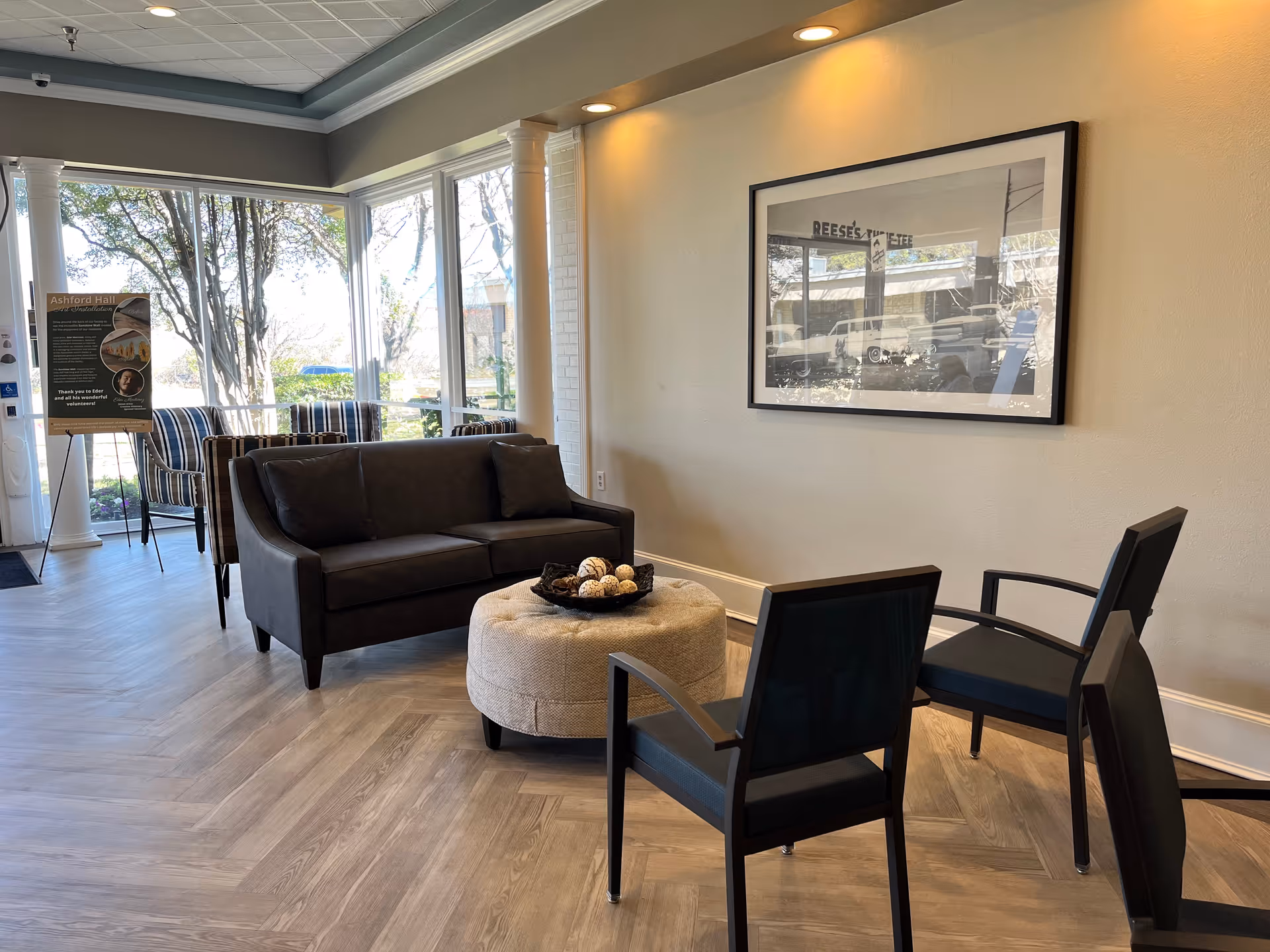 A bright and modern sitting area with a dark gray sofa, two black chairs, and a round beige ottoman with decorative balls on top. Large windows let in natural light, and a framed picture hangs on the wall. A signboard is visible near the entrance with information about Ashford Hall Nursing and Rehabilitation.