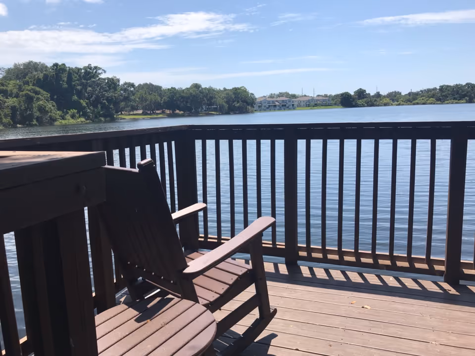 Wooden deck with a wooden chair and table overlooking a large calm lake with trees and buildings in the distance under a partly cloudy sky.