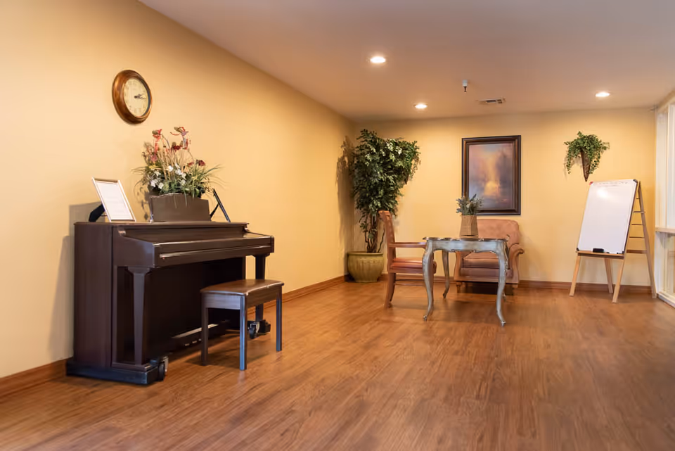 A cozy interior room with wooden flooring featuring a dark brown upright piano with a matching bench on the left side. Above the piano is a round wall clock and a floral arrangement. In the background, there is a small sitting area with a wooden chair, a cushioned armchair, and a decorative table with a plant on it. The walls are painted beige, adorned with a framed painting and a hanging plant. A whiteboard on an easel stands near a window on the right side.