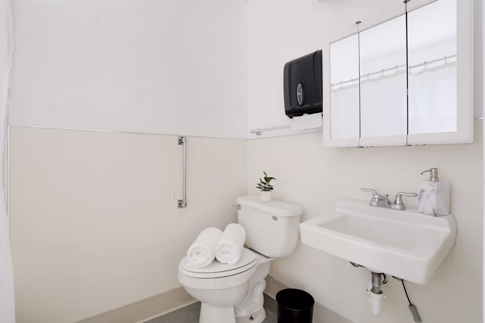 A clean and simple bathroom with a white toilet topped with two rolled white towels and a small potted plant. There is a white sink with a soap dispenser, a mirrored medicine cabinet above it, a black paper towel dispenser on the wall, and a black trash bin on the floor. The walls are painted white and beige with a grab bar installed next to the toilet.