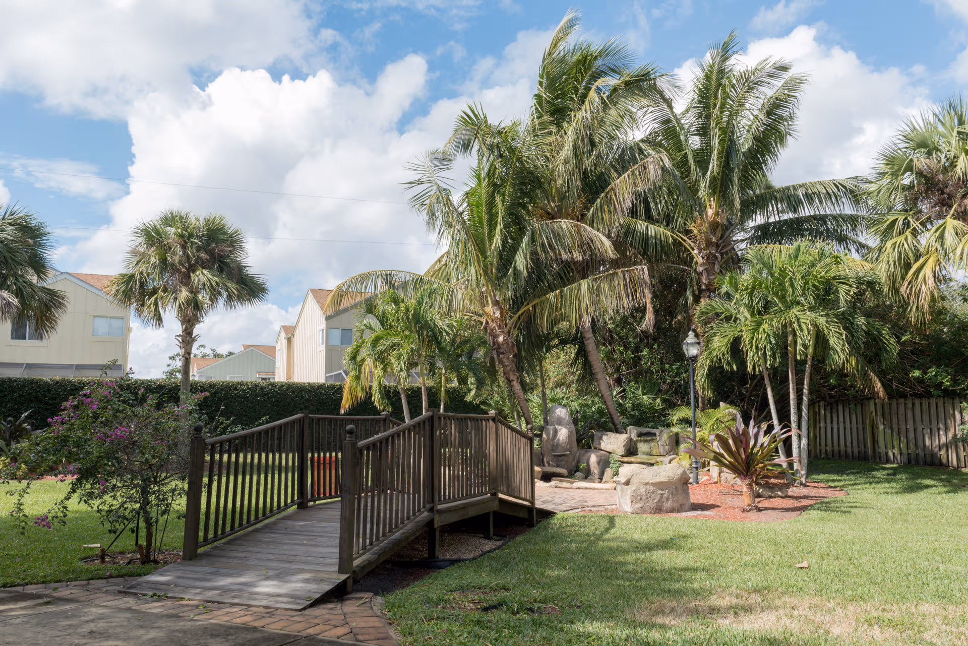 A sunny outdoor garden area with a small wooden bridge over a dry patch, surrounded by green grass, palm trees, and other tropical plants. In the background, there are residential-style buildings with beige siding and red roofs under a partly cloudy sky.