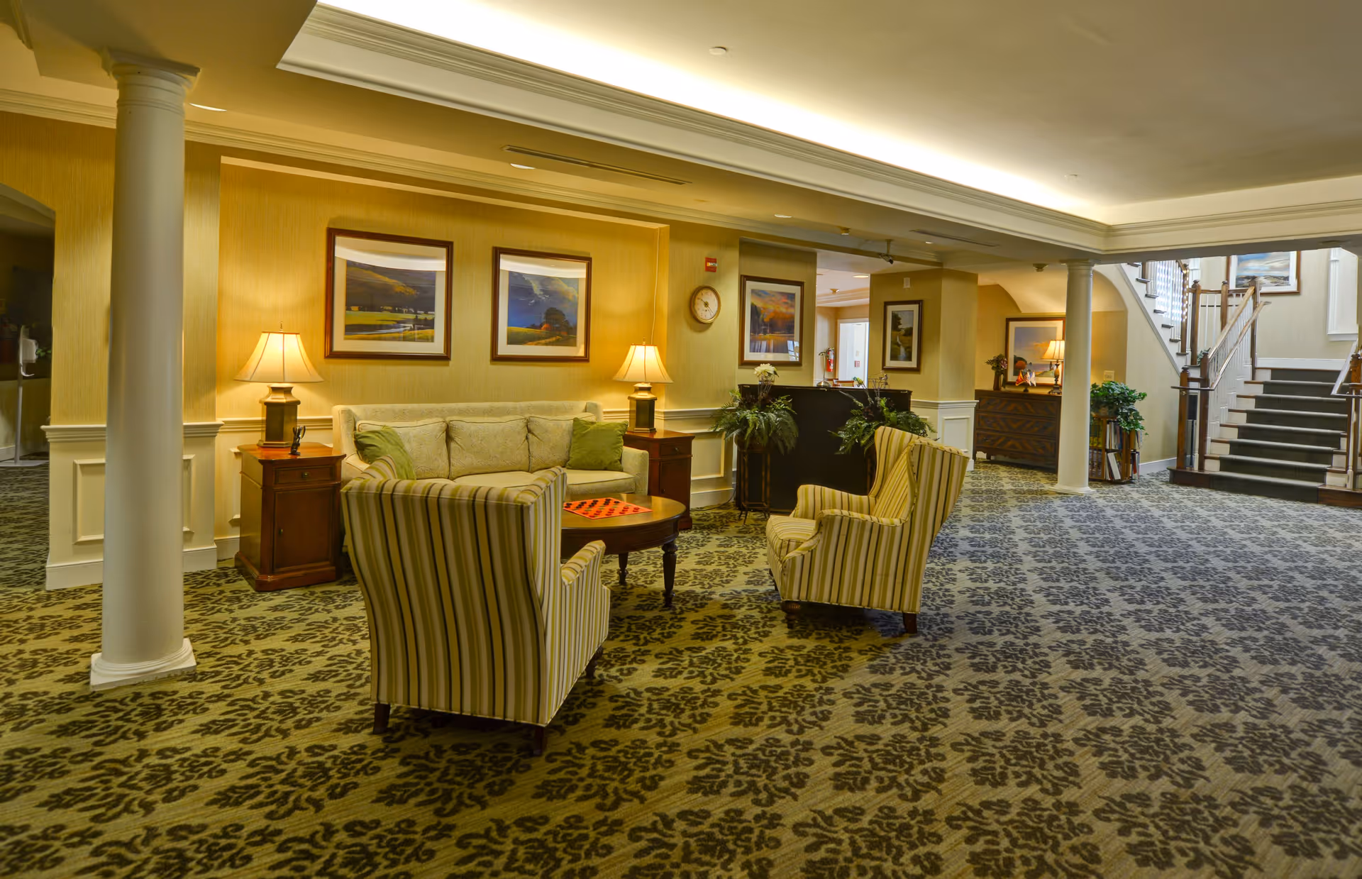 A spacious and warmly lit senior living facility common area with patterned carpet, two striped armchairs, a beige sofa, wooden side tables with lamps, framed landscape paintings on the wall, potted plants, and a staircase leading to an upper floor.