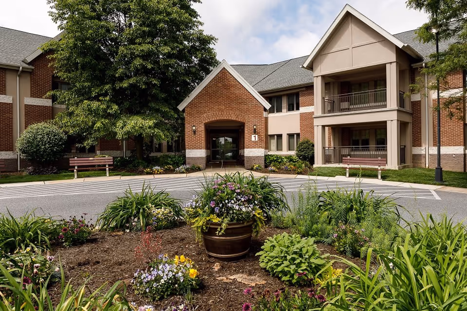 Front exterior view of Phoebe Berks facility showing a brick building with a covered entrance, balconies, benches, and landscaped flower beds with various plants and flowers in the foreground.
