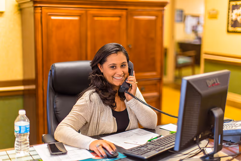 A smiling woman sitting at a desk in an office environment, talking on a corded phone while using a computer mouse. The desk has papers, a water bottle, and a smartphone on it. Behind her is a wooden cabinet and a hallway leading to other rooms.