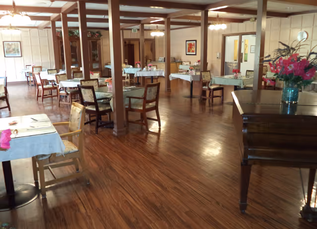 A spacious dining room with wooden floors and several tables covered with light blue tablecloths. Each table has chairs around it and small flower arrangements as centerpieces. The room has wooden beams and columns, framed artwork on the walls, and a piano with a vase of flowers on top. Warm lighting fixtures hang from the ceiling.