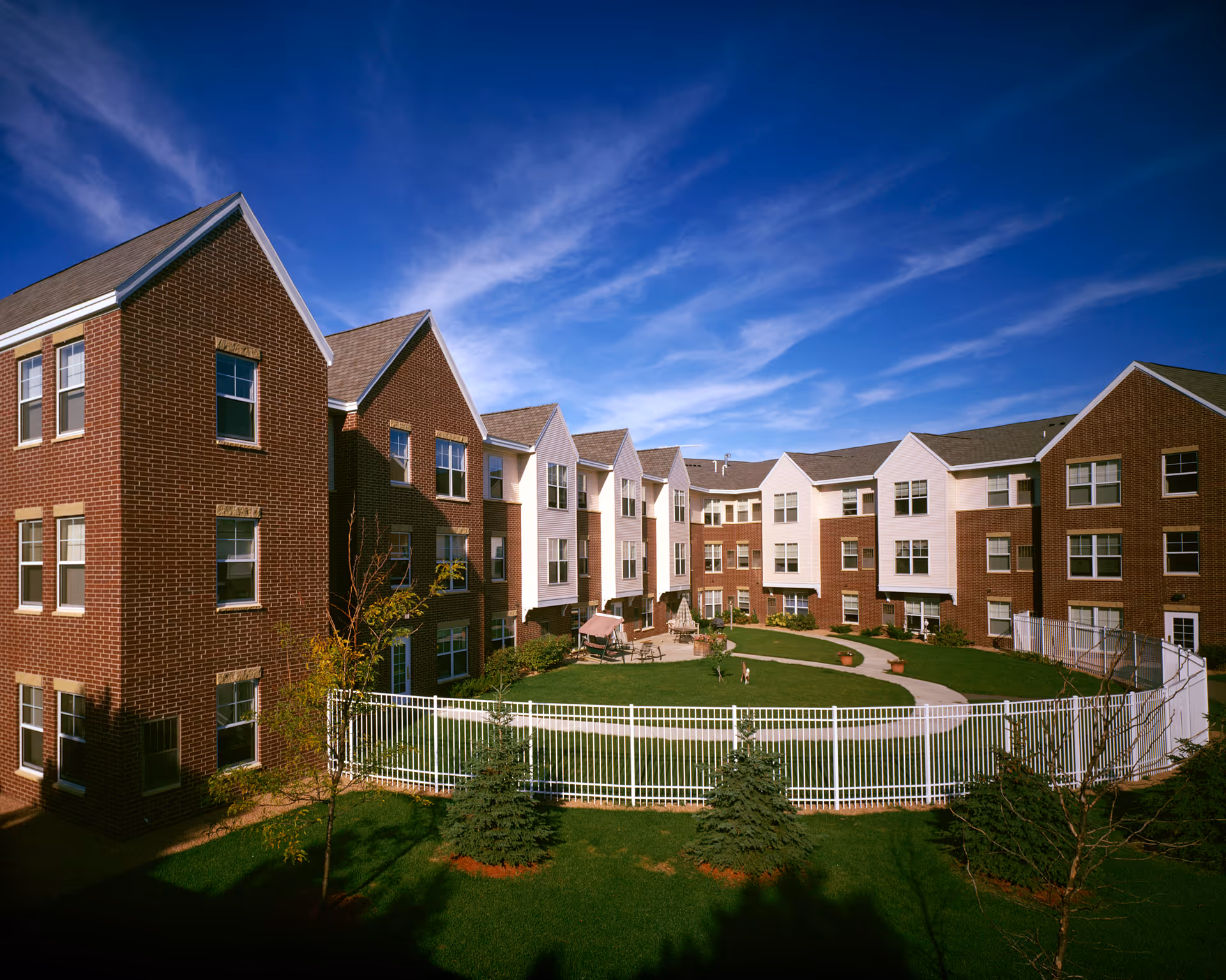 Exterior view of a senior living facility named The Waters of Eden Prairie, showing a three-story brick and white-paneled building surrounding a fenced green courtyard with a paved walkway, small trees, and outdoor seating under a clear blue sky.