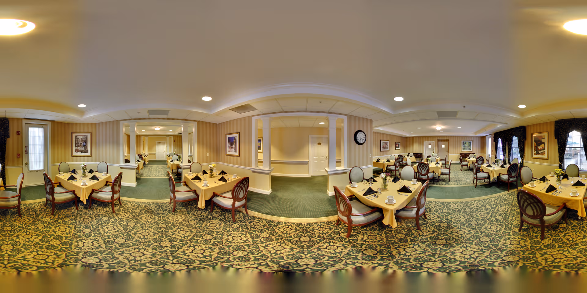 A spacious dining room in a senior living facility with multiple tables covered in yellow tablecloths, each set with black napkins, cups, and small flower arrangements. The room has patterned green carpet, beige walls with framed artwork, and several windows with dark curtains allowing natural light to enter. Chairs with wooden frames and cushioned seats surround the tables.
