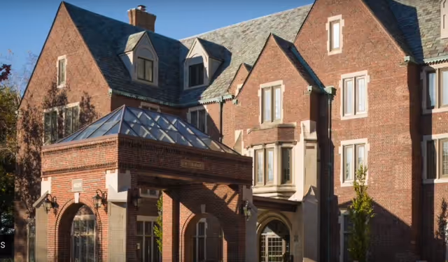 Exterior front facade of a brick, multi-story senior living building with gabled roofs, arched entrance, and a glass-roofed portico.