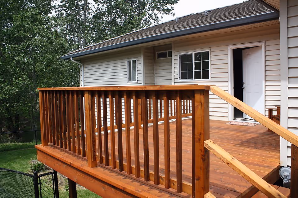 Wooden deck attached to a beige house with a white door and window, surrounded by green trees and grass.