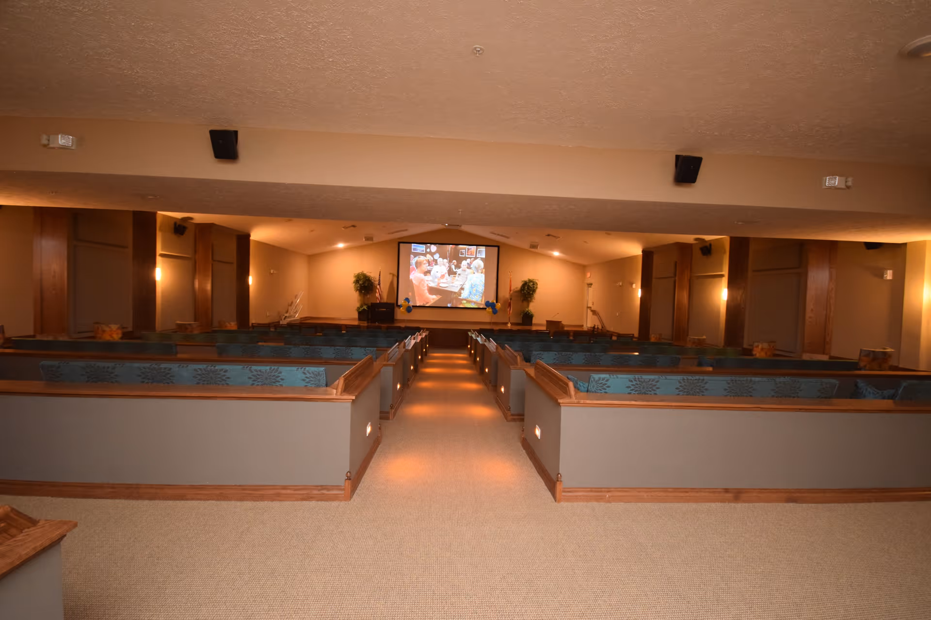 Interior view of a large room with rows of wooden pew-style seating facing a screen at the front displaying a group of people. The room has beige walls, carpeted floor, and soft lighting along the aisle and walls.