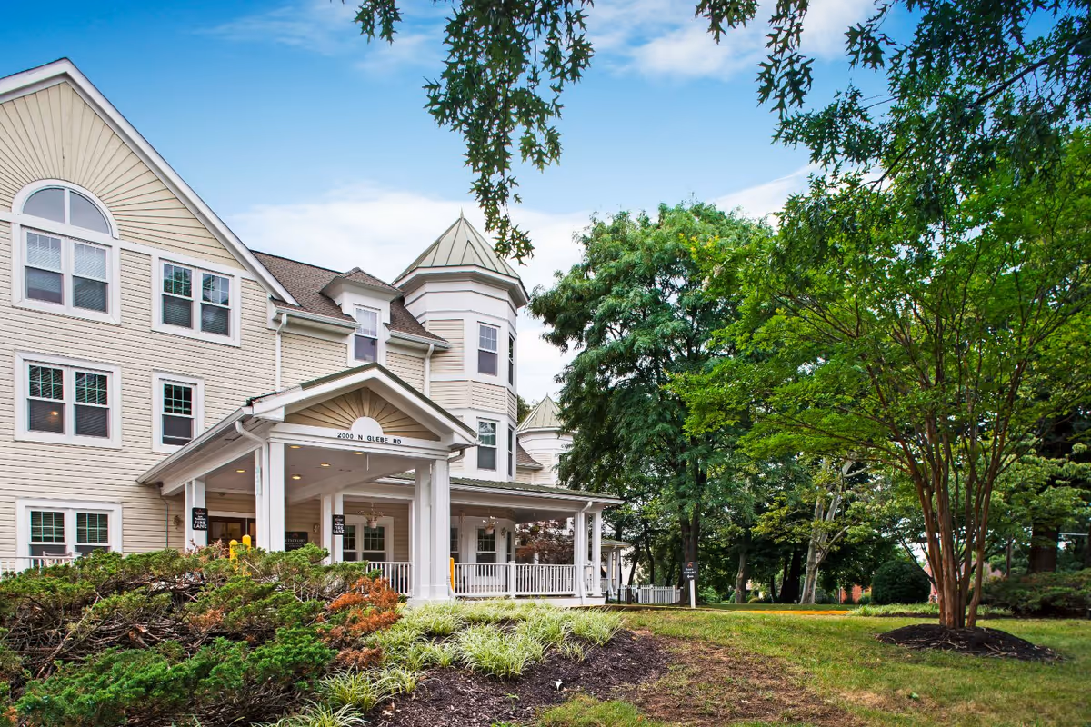 Exterior view of a multi-story senior living facility building with beige siding and white trim. The building features a covered entrance with white columns and a sign displaying the address 2000 N Glebe Rd. The surrounding area includes well-maintained landscaping with green shrubs, trees, and a grassy lawn under a partly cloudy blue sky.