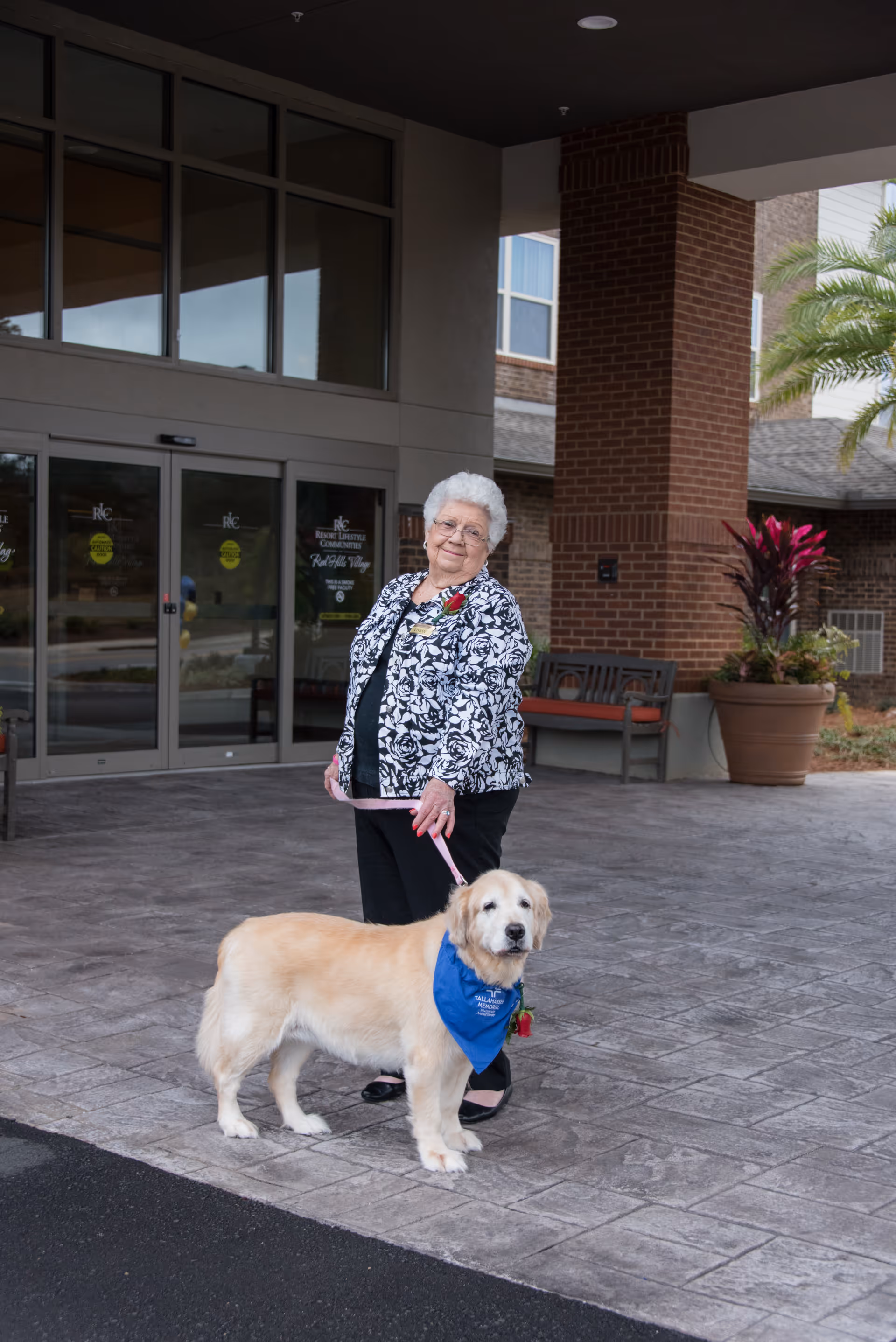 An elderly woman smiles while holding a leash attached to a golden retriever at the covered entrance of a retirement residence.