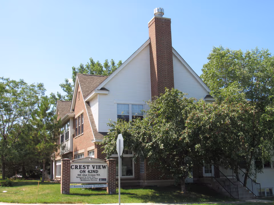 Exterior view of a two-story brick and white siding building with a chimney, surrounded by trees and greenery under a clear blue sky. A sign in front reads 'Crest View On 42nd' with additional information about assisted living availability.