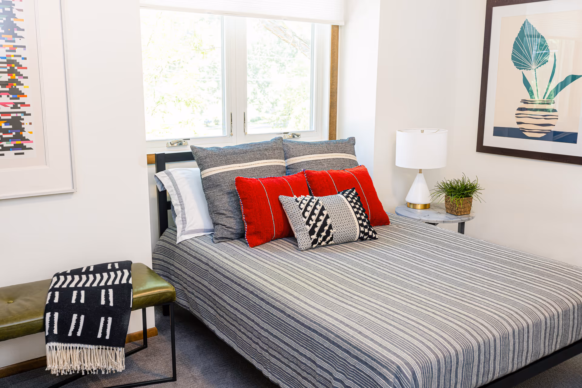 A neatly made bed with a gray striped bedspread and multiple pillows in gray, red, and black-and-white patterns. The bed is positioned under a window with white blinds. To the right of the bed is a small round side table with a white lamp and a small potted plant. On the left side, there is a green bench with a black and white patterned throw blanket draped over it. The walls are white and decorated with framed artwork.