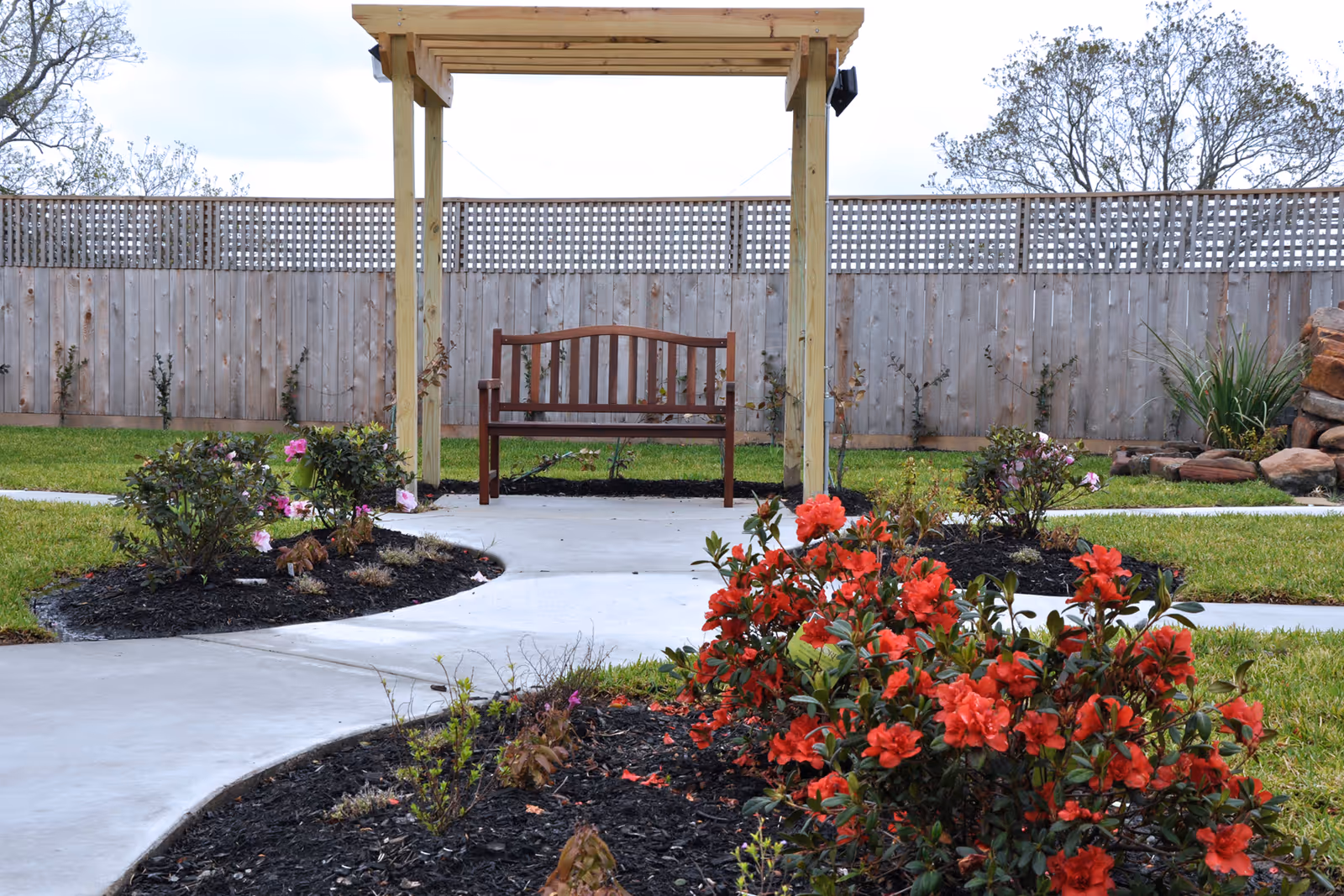 A peaceful outdoor garden area with a wooden bench under a wooden pergola. The garden features a concrete pathway winding through flower beds with red and pink blooming flowers, surrounded by green grass and a wooden fence in the background.