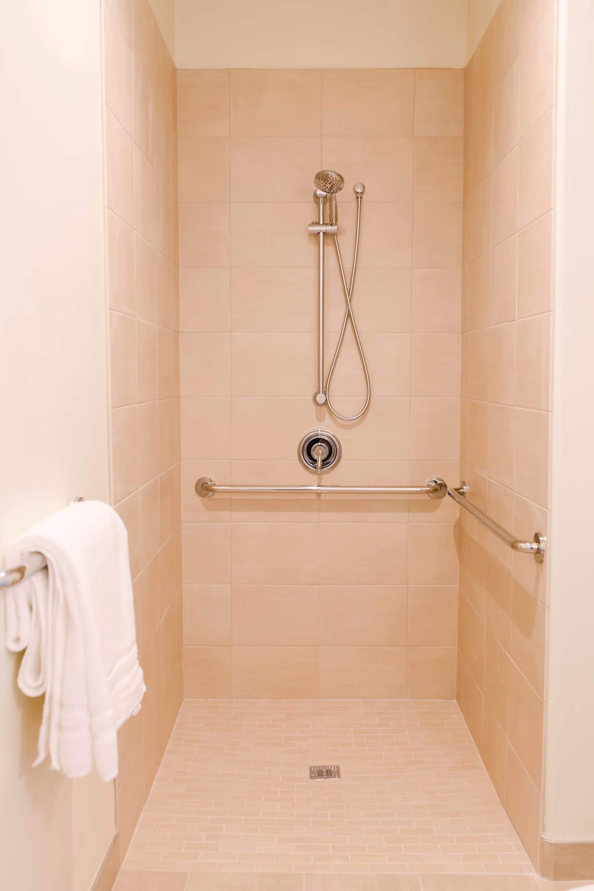 A beige tiled walk-in shower with a handheld showerhead mounted on the wall, two horizontal grab bars on the side walls, and a white towel hanging on a towel bar outside the shower.