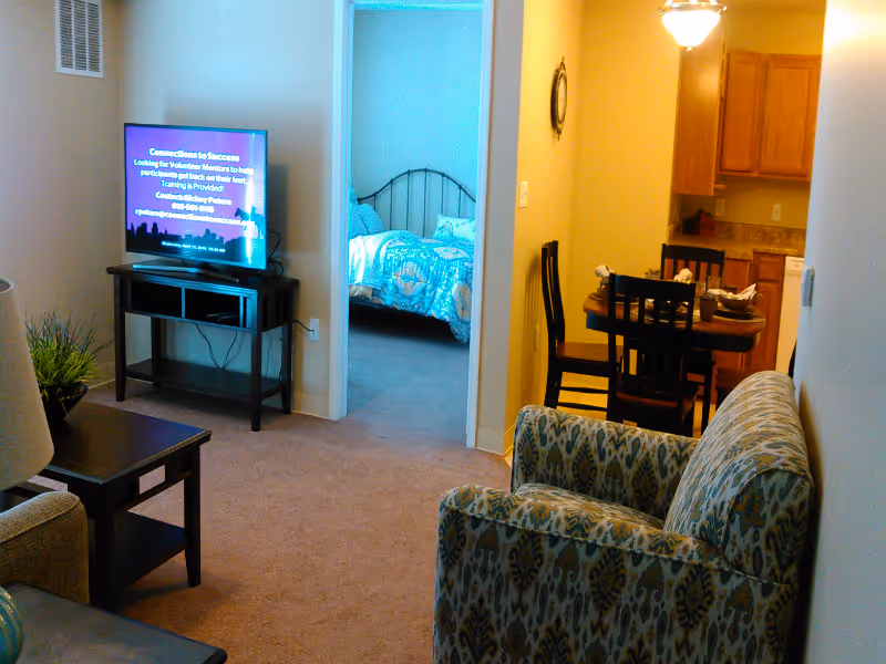 Living room with a patterned armchair, TV on a stand, coffee table, and a view into a bedroom and dining area.