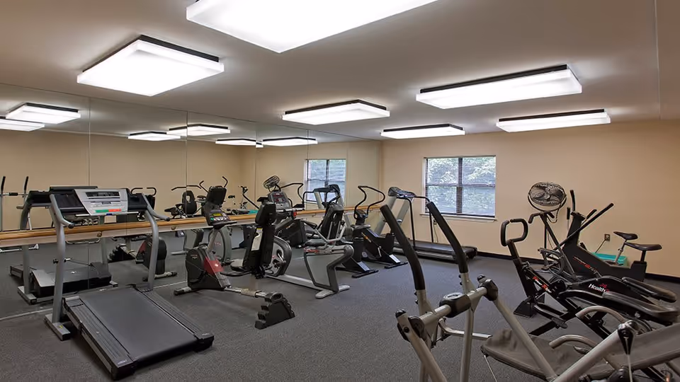 A fitness room with various exercise equipment including treadmills, stationary bikes, and elliptical machines. The room has beige walls, a large mirror covering one wall, two windows letting in natural light, and ceiling lights providing bright illumination.