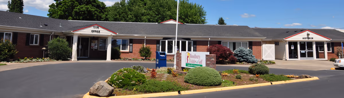 Exterior view of a single-story brick building with a sign in front that reads 'Woodburn Estates & Golf Active 55+ Community.' The building has two labeled entrances: one marked 'Office' and the other 'Auditorium.' There is a landscaped area with bushes and a mailbox in front, and a clear blue sky with some clouds above.