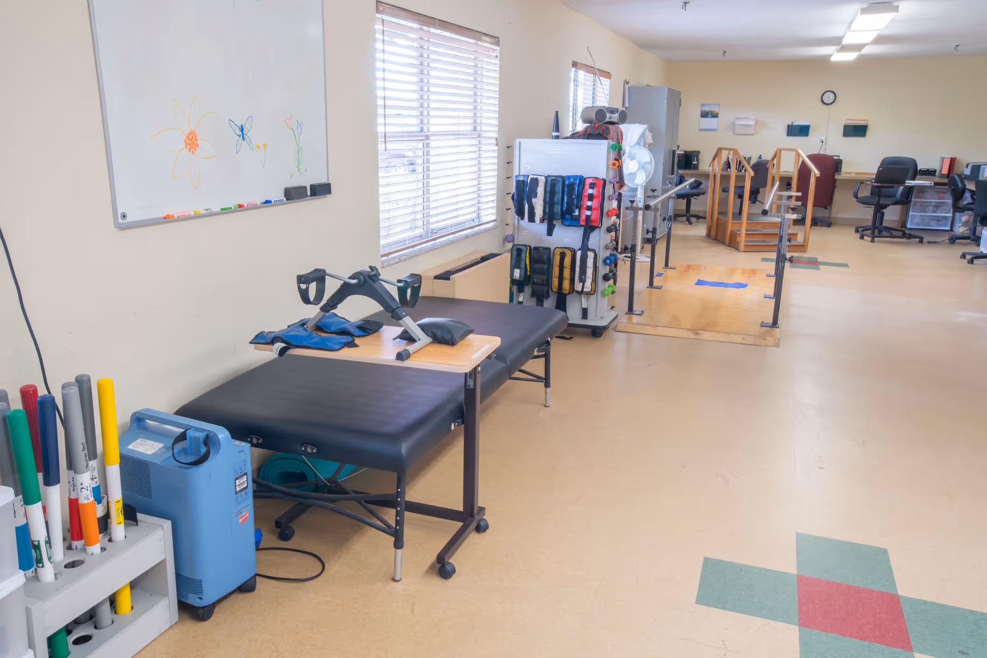 A physical therapy room with exercise equipment including a treatment table, hand weights, parallel bars for walking practice, and various therapy tools organized on a rack. The room has a window with blinds, a whiteboard with simple flower drawings, and office chairs and desks in the background.
