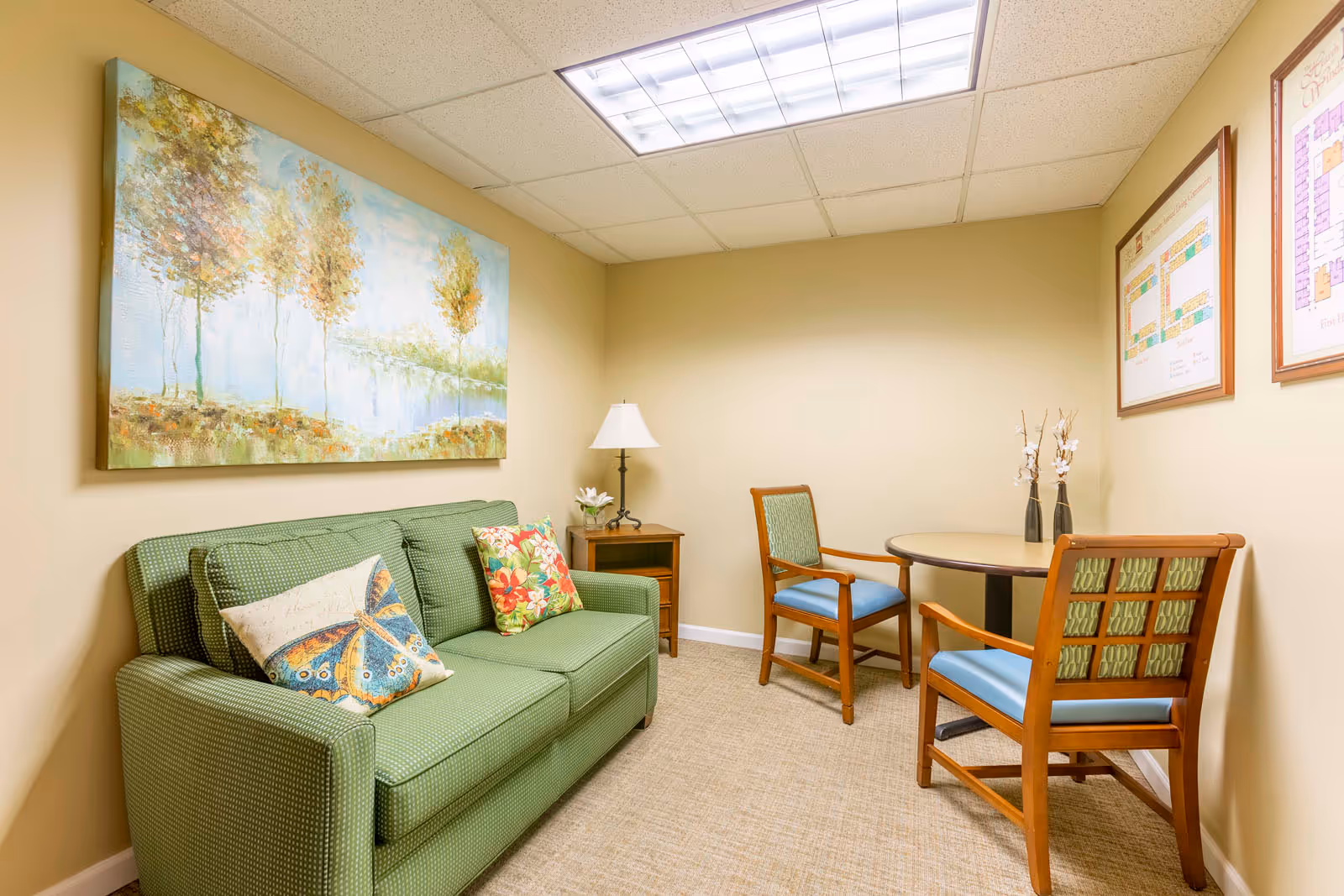 A small, cozy room with a green upholstered sofa adorned with decorative pillows, a wooden side table with a lamp and flower vase, two wooden chairs with blue cushions around a round table, and framed floor plans on the wall. A large painting of trees and a lake hangs above the sofa.