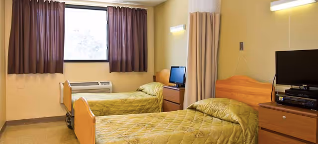 A simple bedroom with two single beds covered in green quilts, each with a wooden headboard. Between the beds is a small wooden nightstand with a flat-screen TV and electronic devices on top. A window with brown curtains is on the far wall, and a beige privacy curtain hangs between the beds. The walls are painted light yellow.