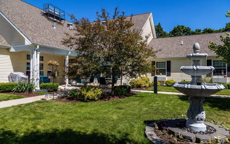Outdoor courtyard area of a senior living facility with a three-tiered stone fountain in the foreground, green grass, a tree, and a beige building with white trim and a covered porch in the background under a clear blue sky.