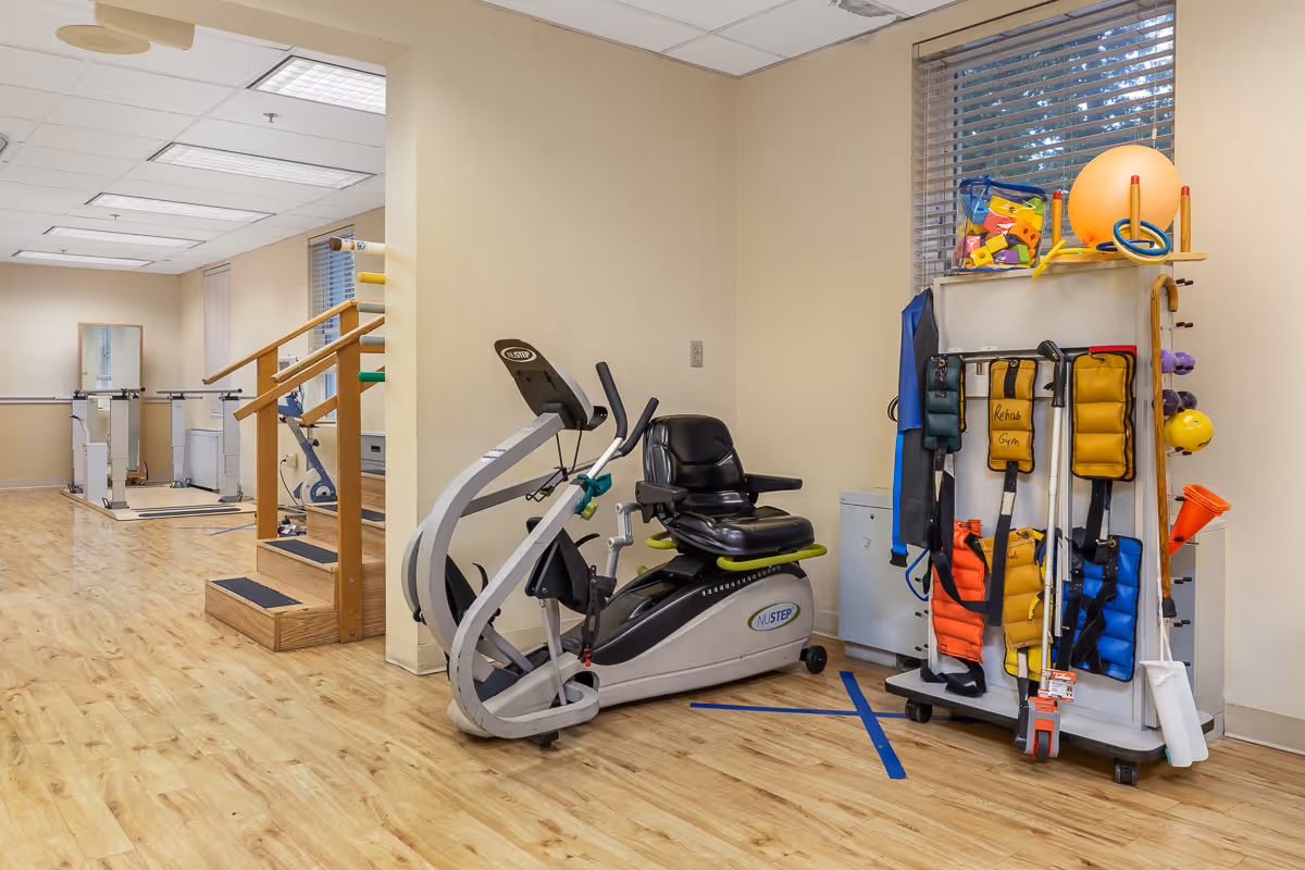 A rehabilitation gym area with exercise equipment including a NuStep recumbent cross trainer, parallel bars for walking exercises, and a rack holding various weighted vests, therapy balls, and other physical therapy tools. The room has light wood flooring, beige walls, and a window with blinds.