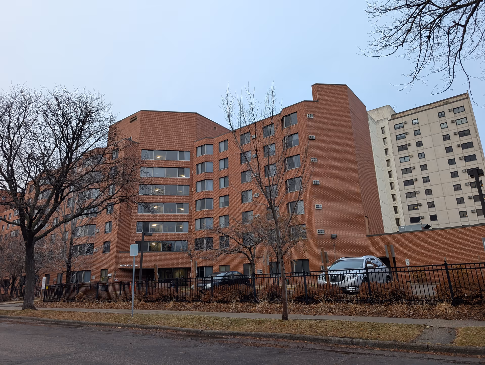 Exterior view of a multi-story brick apartment building with several windows and a fenced area in front. Leafless trees and parked cars are visible along the street and inside the fenced area. The sky is overcast.