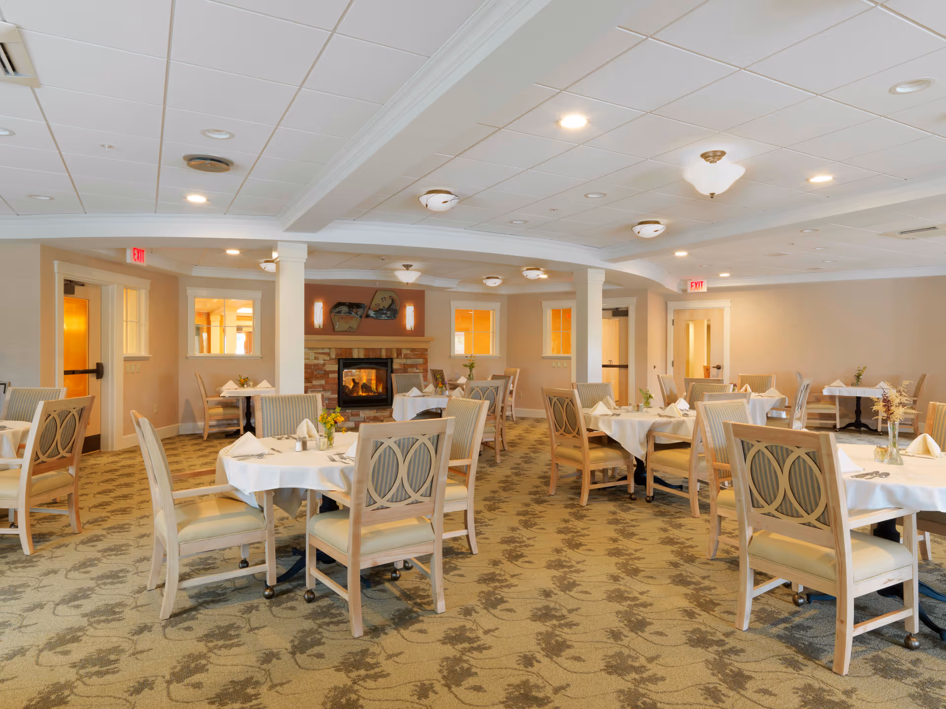 Well-lit dining room with round tables set for meals, wooden chairs, and a fireplace in the background.