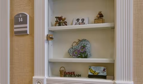 A white built-in wall cabinet with three shelves displaying decorative items including small figurines, a heart-shaped ornament with floral design, and a small basket. To the left of the cabinet is a beige wall with a room number plaque labeled '14'.