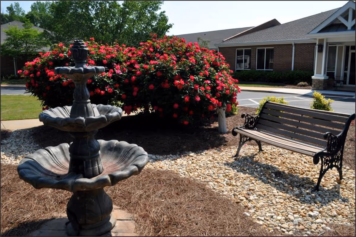 Outdoor garden area with a multi-tiered stone fountain, a wooden bench with decorative metal armrests, vibrant red flowering bushes, and a brick building in the background under a clear sky.
