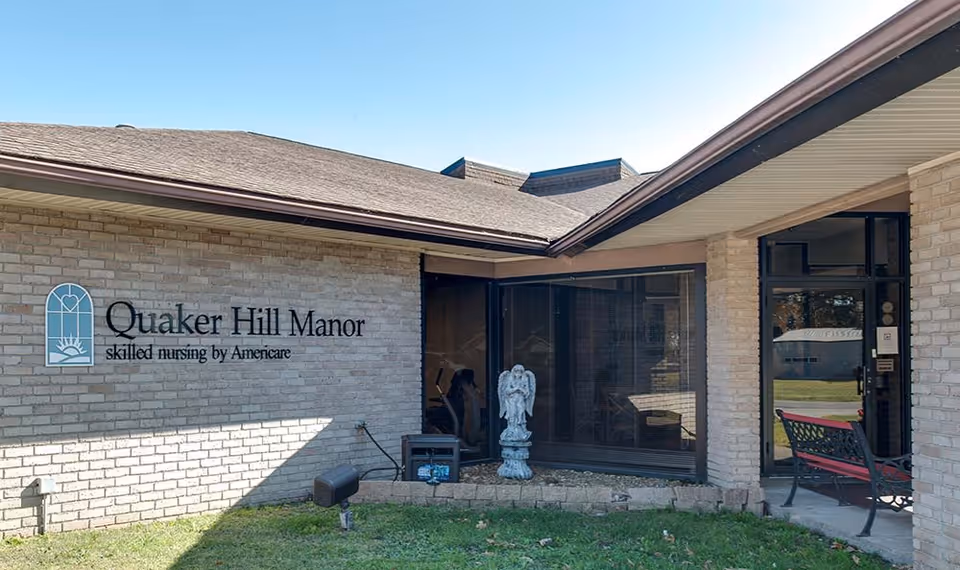 Exterior view of Quaker Hill Manor, a skilled nursing facility by Americare, showing a brick building with a sign on the wall, a small statue of an angel, a bench, and a glass door entrance under a clear blue sky.