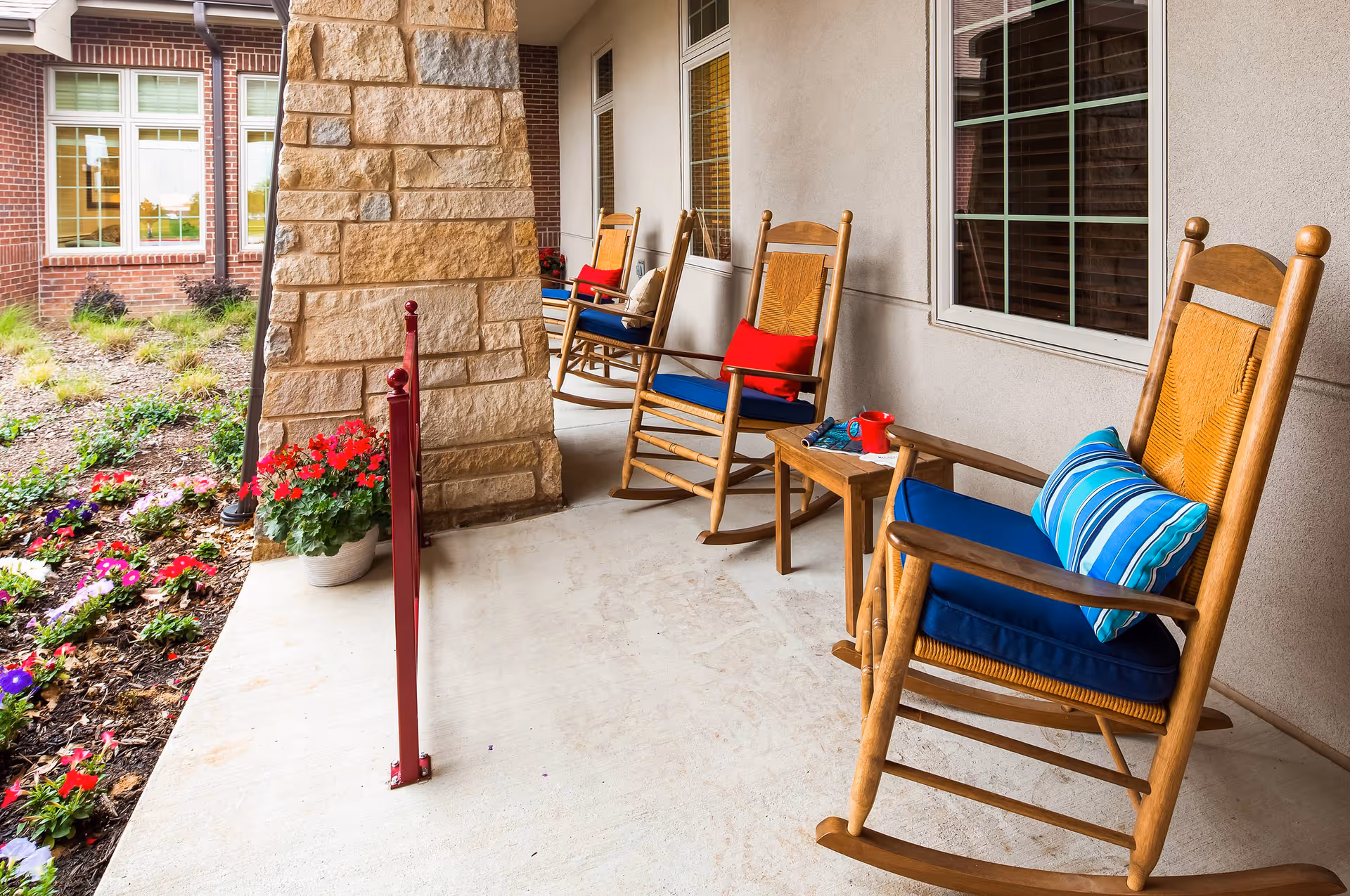 A covered outdoor porch area with wooden rocking chairs featuring blue cushions and colorful pillows. There is a small wooden table with a red mug and some reading materials. The porch overlooks a garden bed with various flowers and plants, and the building exterior is made of brick and stone with large windows.