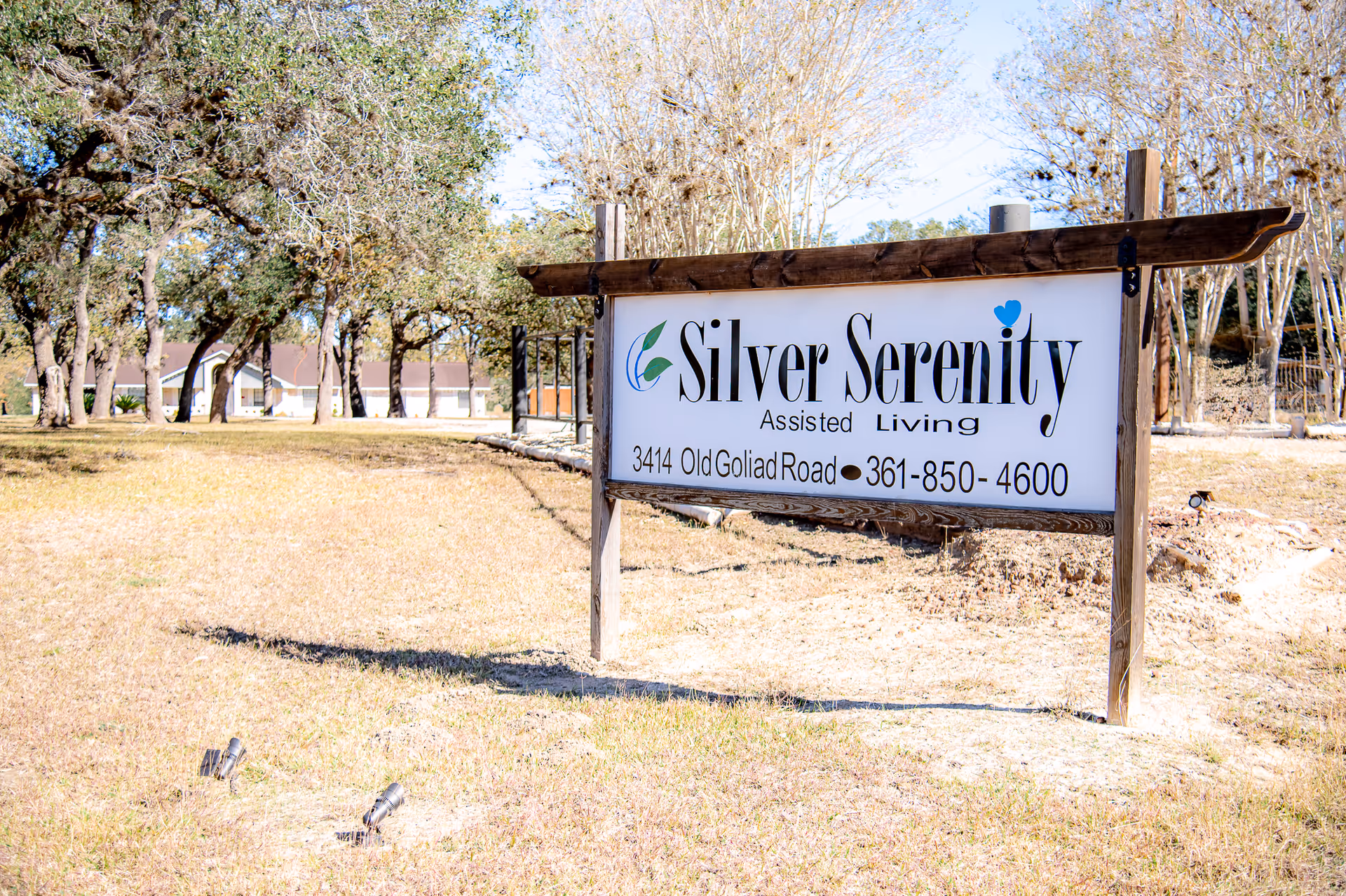Outdoor view of a wooden sign for Silver Serenity Assisted Living located on a grassy area with trees and buildings in the background under a clear sky.