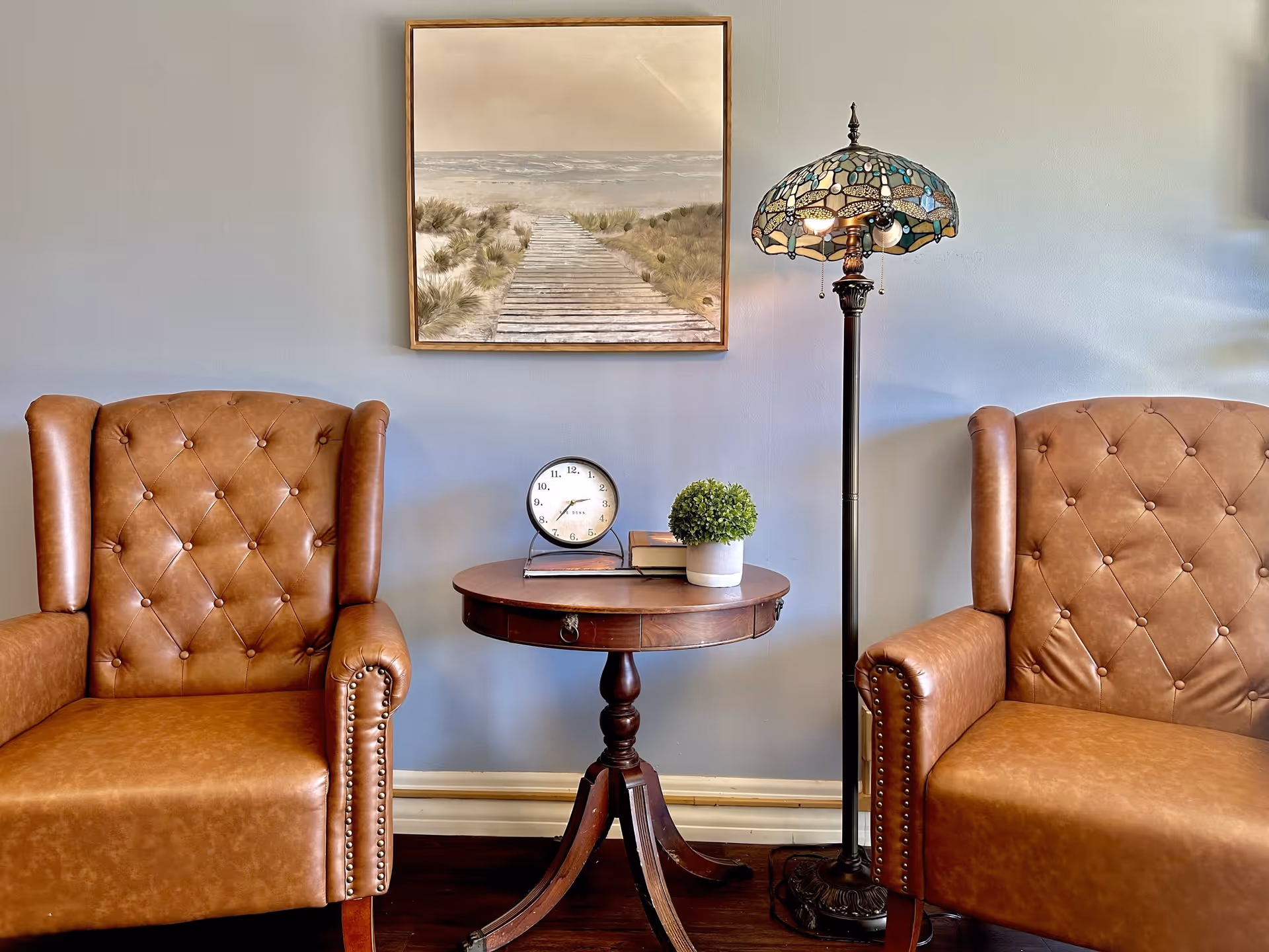 Two brown leather armchairs with tufted backs are positioned on either side of a round wooden side table. On the table, there is a small round clock, a stack of books, and a small potted plant. A tall floor lamp with a stained glass shade stands to the right of the table. Above the table hangs a framed painting of a wooden boardwalk leading through sand dunes to the ocean.