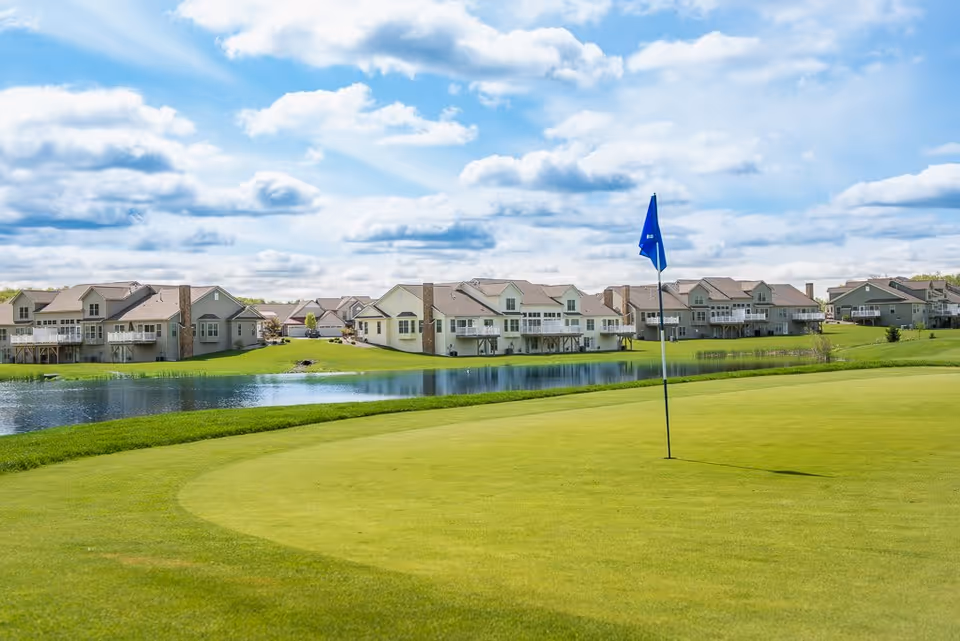 A scenic view of a golf course green with a blue flagstick in the foreground, a pond behind it, and a row of residential buildings in the background under a partly cloudy sky.