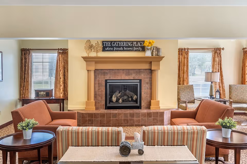 Cozy communal living room with a central fireplace, upholstered chairs and tables, and a sign above the mantel reading "The Gathering Place".