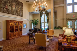 Spacious lounge with upholstered chairs and sofas arranged around small tables, a grand piano, a large chandelier, a tapestry on the wall, and tall windows.