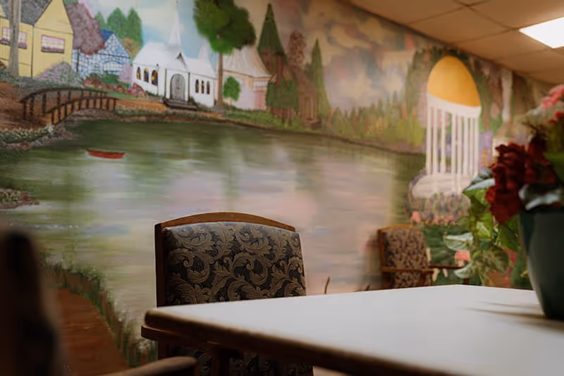 A small dining table with upholstered chairs in a room featuring a large lakeside mural and a floral centerpiece.