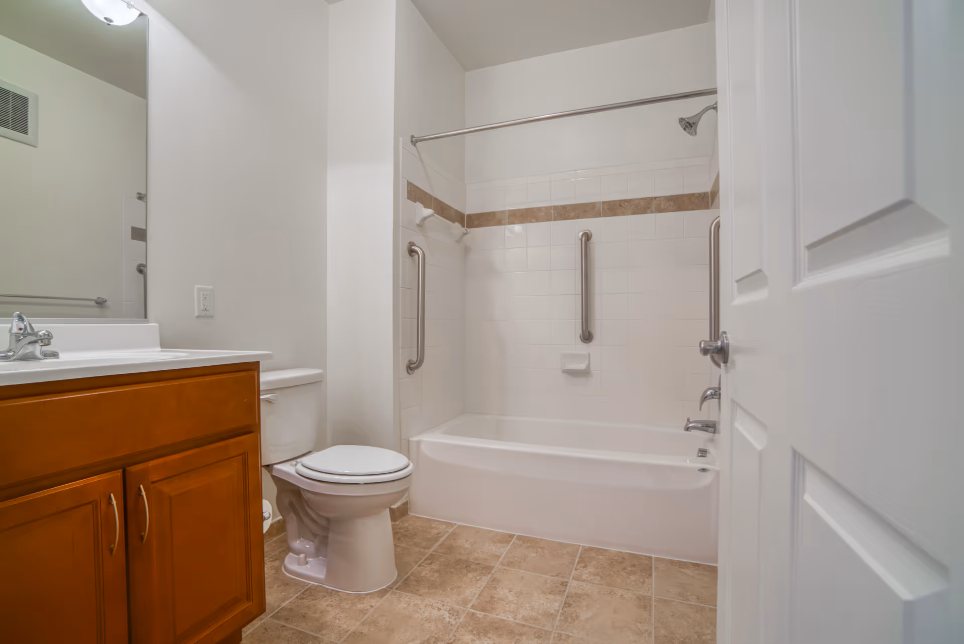 A clean bathroom with a white bathtub and shower combination featuring grab bars, a white toilet, a wooden vanity with a sink and faucet, a large mirror, and beige tiled floor.