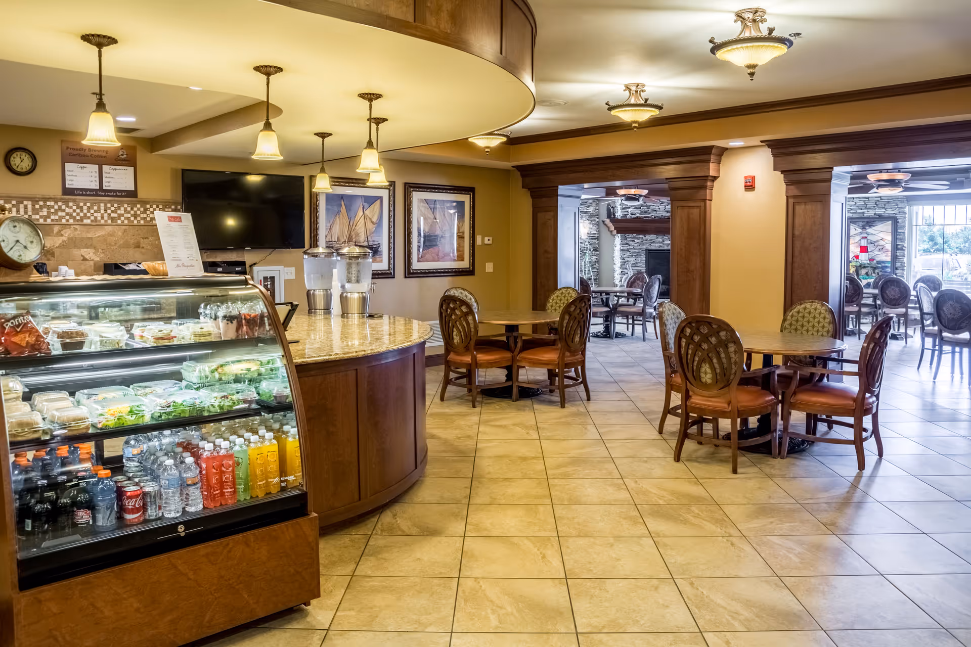 Interior view of a senior living facility dining area with a curved wooden counter displaying bottled drinks and packaged food items. The room has tiled floors, round tables with chairs, pendant lights hanging from the ceiling, and framed artwork on the walls. In the background, there is a seating area with more tables and chairs near large windows.