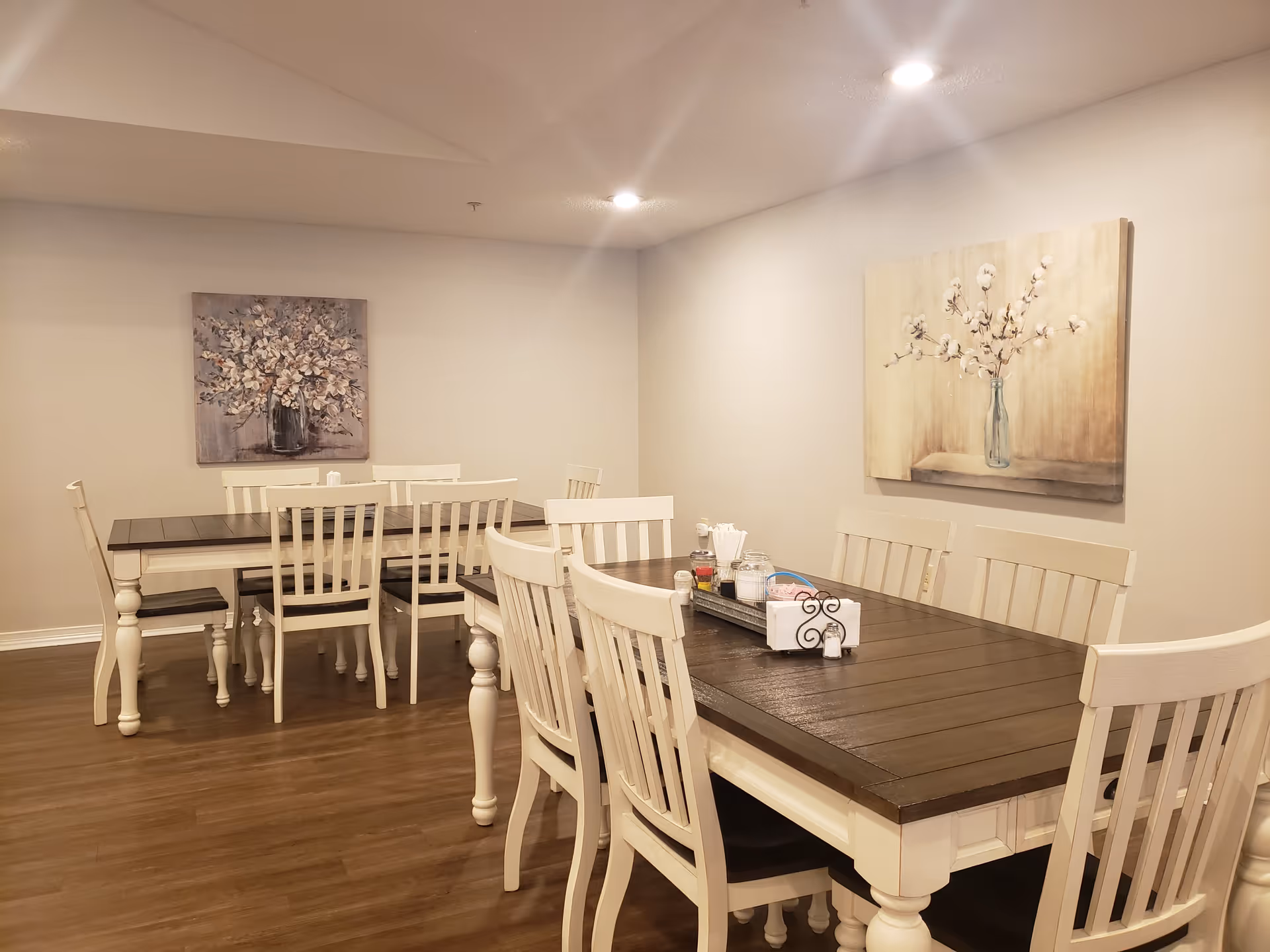 A dining room with two wooden tables and multiple white chairs. The tables have dark brown tops and white legs. On the table in the foreground, there is a tray with condiments and napkins. The walls are light-colored and decorated with two paintings of flowers in vases. The floor is wooden, and recessed ceiling lights illuminate the room.
