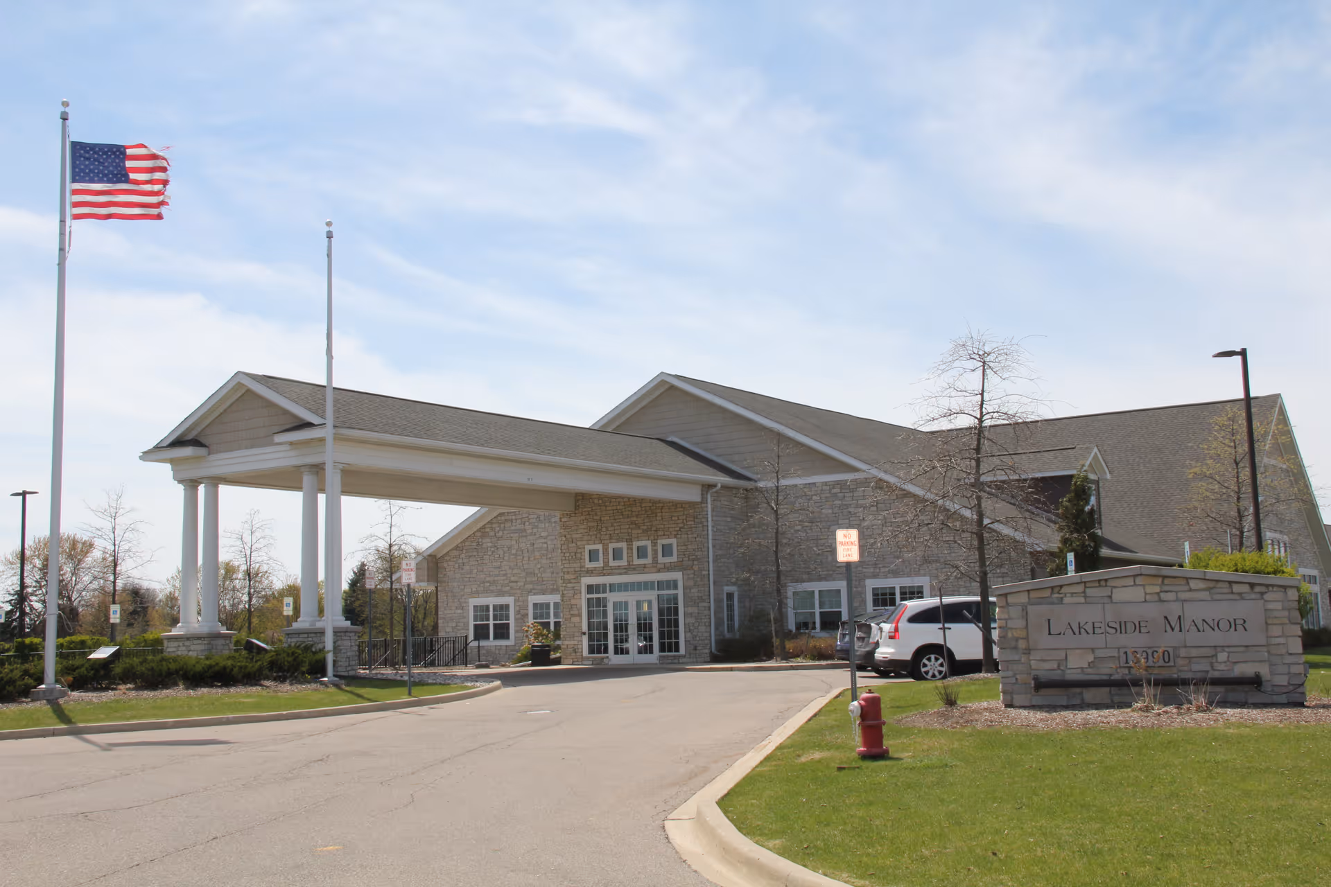 Exterior view of Lakeside Manor facility showing a stone building with a covered entrance, an American flag on a flagpole, a parking area with cars, and a stone sign with the facility name and address.