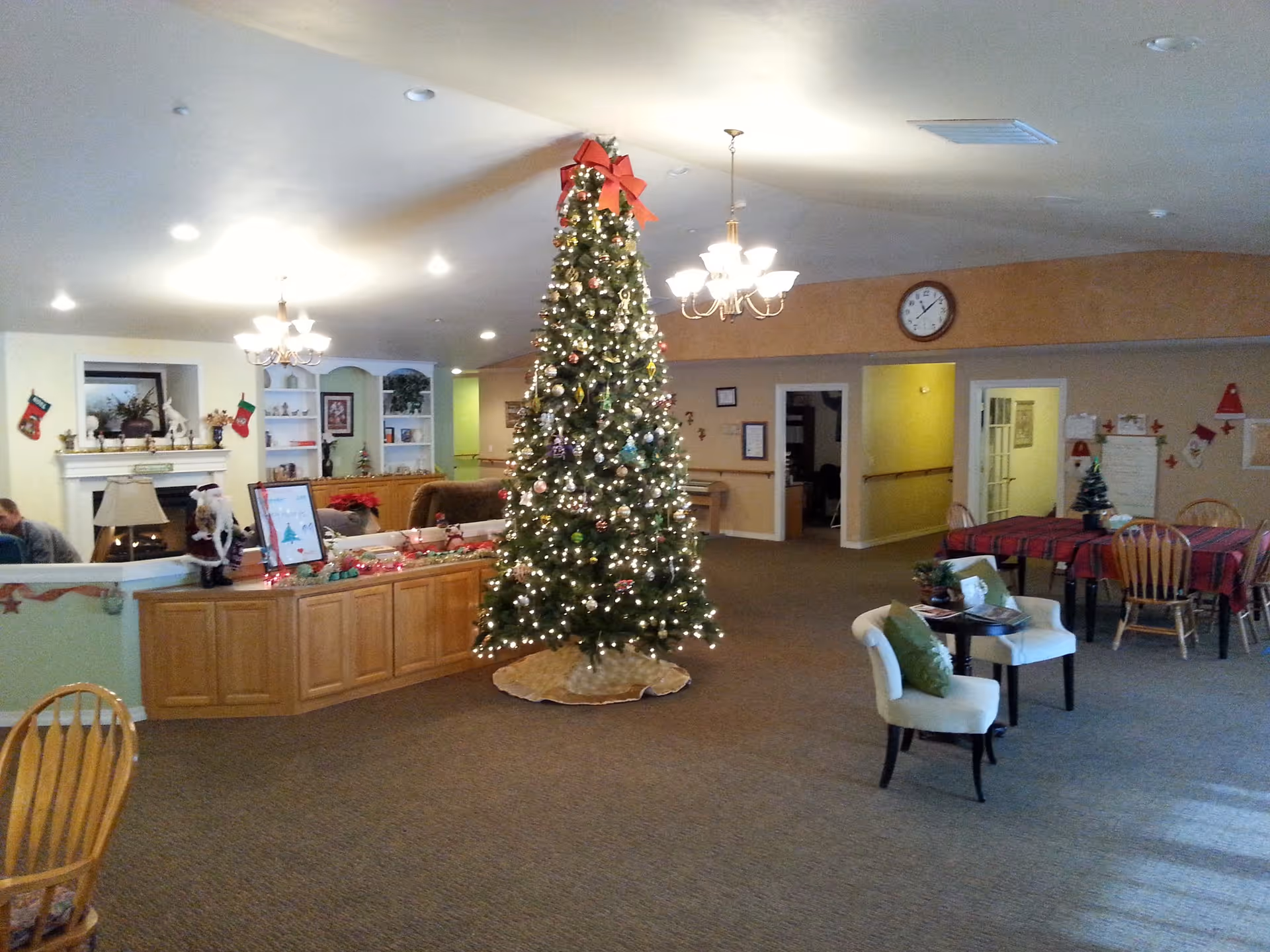 A spacious living room area decorated for Christmas with a tall Christmas tree adorned with lights and ornaments in the center. The room features carpeted floors, wooden furniture including chairs and tables, a fireplace with stockings hanging, and festive decorations throughout. There are chandeliers hanging from the ceiling and a clock on the wall.